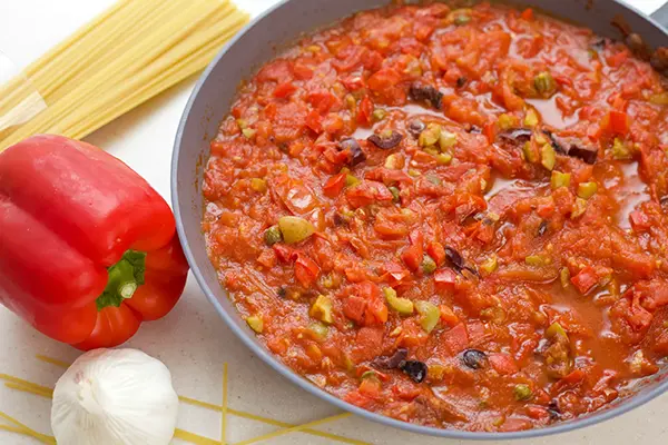 Puttanesca sauce ready in a skillet, surrounded by one garlic heal, one red bell pepper and spaghetti pasta